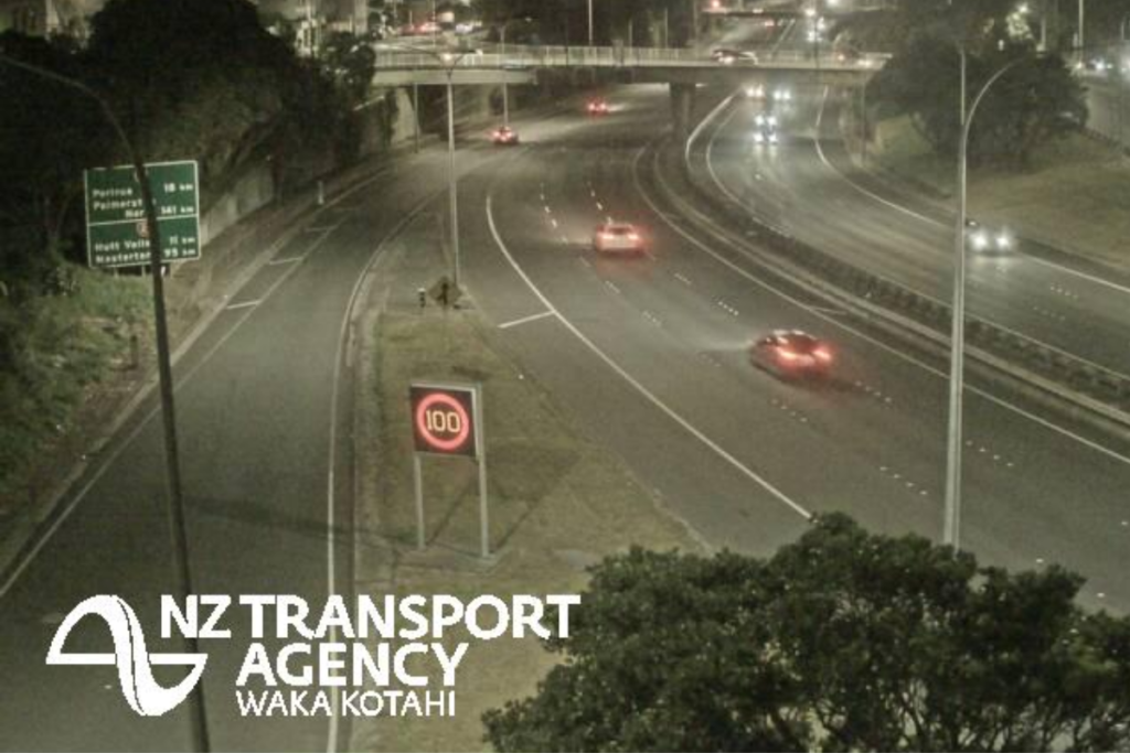 Night-time traffic travelling on Wellington’s urban motorway beneath an overbridge, with illuminated road signs and a 100km/h speed display.