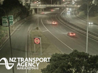 Night-time traffic travelling on Wellington’s urban motorway beneath an overbridge, with illuminated road signs and a 100km/h speed display.