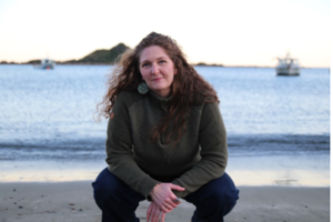 Wellington-based Norwegian songwriter Hanne Jøstensen crouches on a beach with the sea and boats behind her.