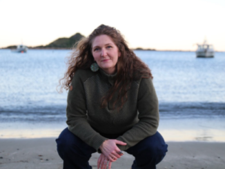 Wellington-based Norwegian songwriter Hanne Jøstensen crouches on a beach with the sea and boats behind her.