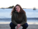 Wellington-based Norwegian songwriter Hanne Jøstensen crouches on a beach with the sea and boats behind her.