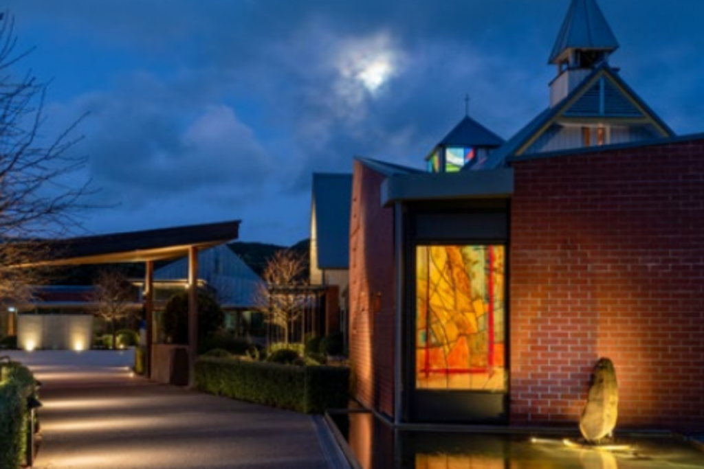 Our Lady’s Home of Compassion in Island Bay at dusk, with an illuminated stained-glass artwork by John Drawbridge visible in the chapel.