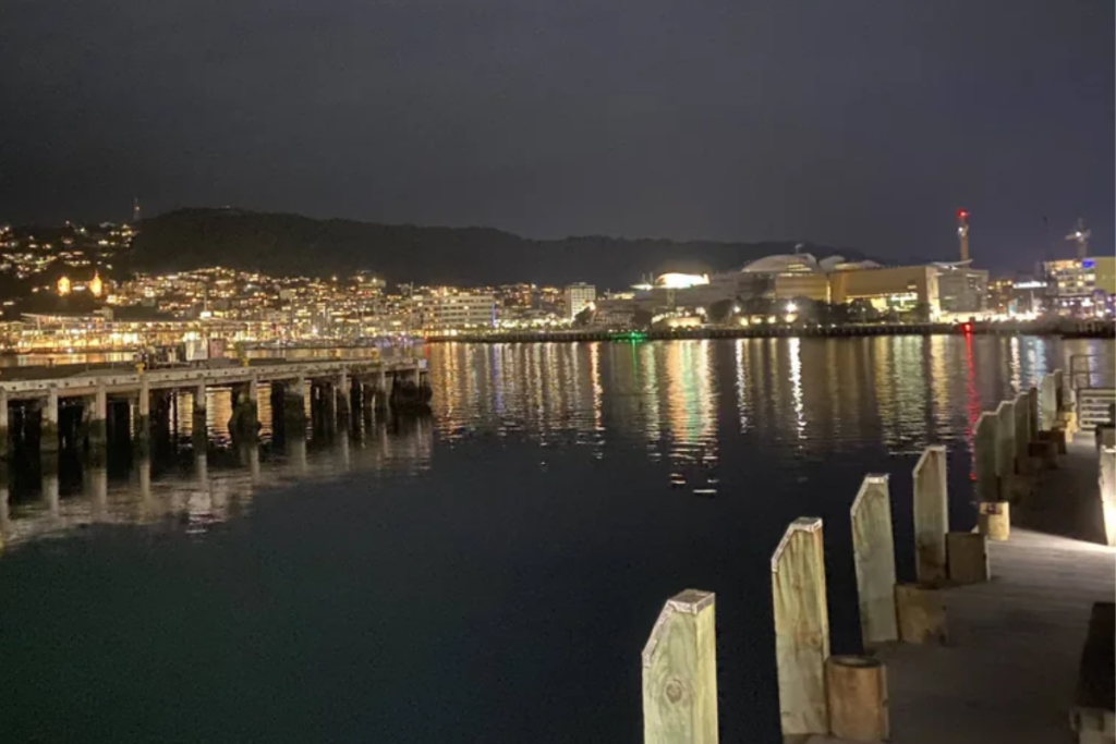 Wellington Harbour at night, with waterfront lights reflecting across the water and the city hills in the background.