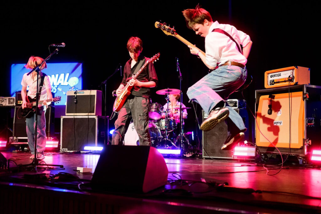 Four young members of Cavemen perform on stage with guitars, bass and drums under concert lighting.