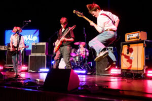 Four young members of Cavemen perform on stage with guitars, bass and drums under concert lighting.