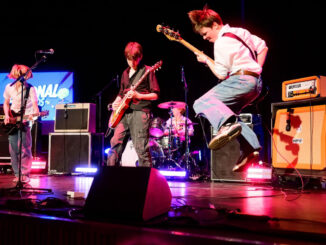 Four young members of Cavemen perform on stage with guitars, bass and drums under concert lighting.