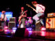 Four young members of Cavemen perform on stage with guitars, bass and drums under concert lighting.