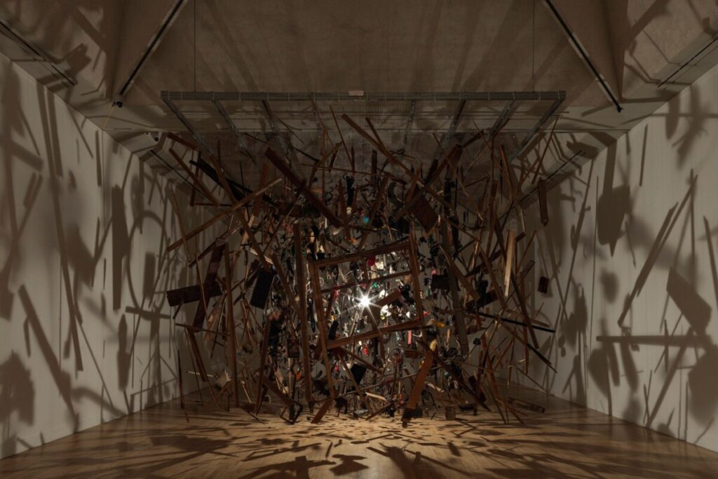 The remains of a garden shed suspended around a light in a gallery room, with the fragments casting long shadows across the walls.
