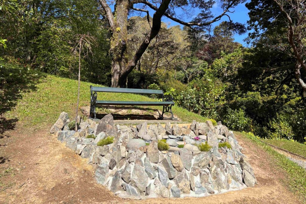 A green park bench behind a circular stone garden beneath leafy trees.