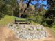 A green park bench behind a circular stone garden beneath leafy trees.