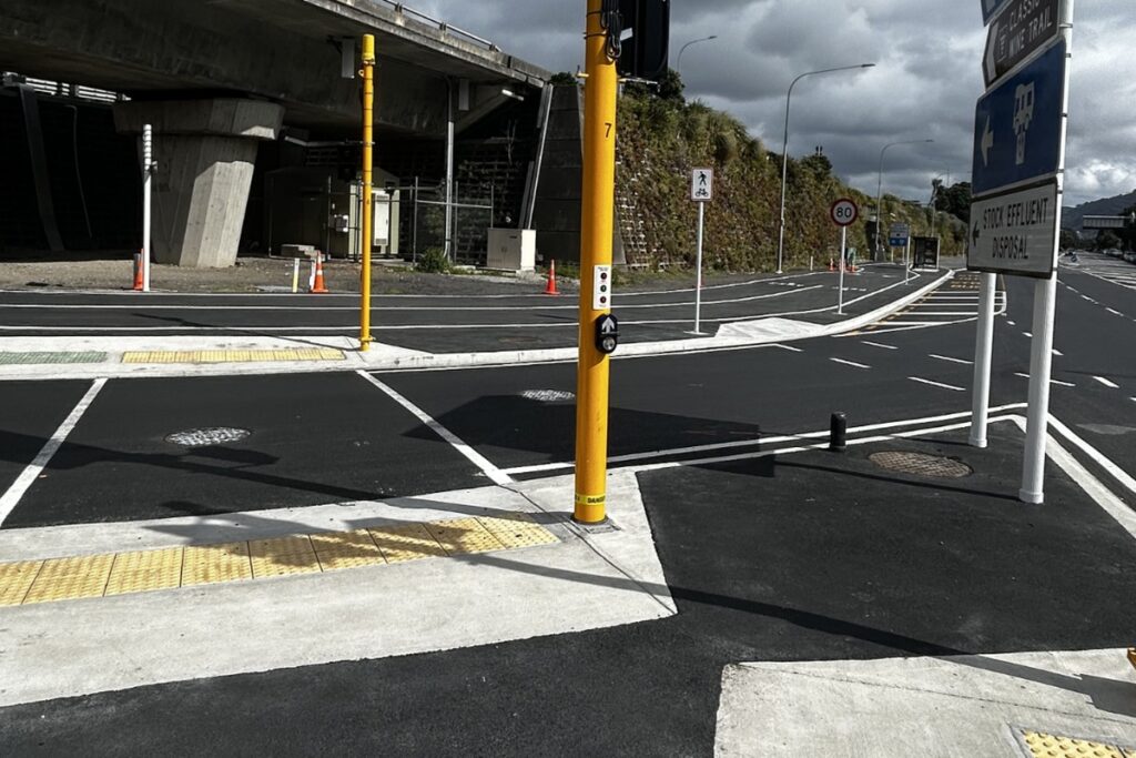 A signal-controlled crossing with yellow poles and tactile paving on Hutt Road beside a concrete viaduct.