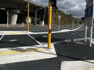 A signal-controlled crossing with yellow poles and tactile paving on Hutt Road beside a concrete viaduct.