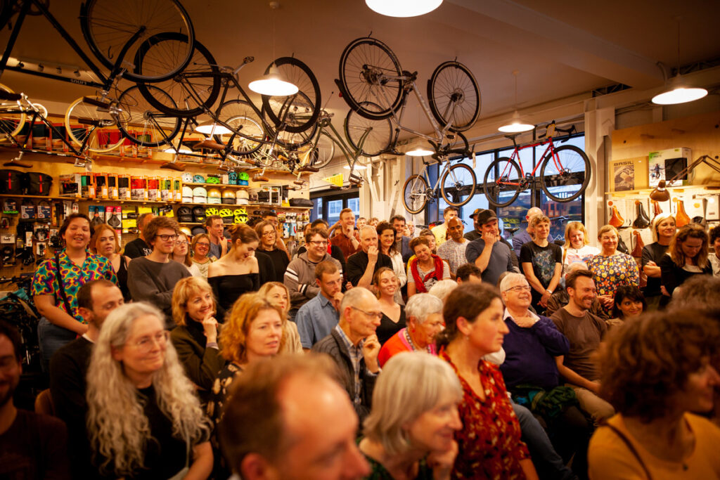 An audience gathered inside a bicycle shop with bicycles hanging overhead.