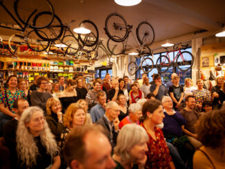 An audience gathered inside a bicycle shop with bicycles hanging overhead.