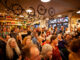 An audience gathered inside a bicycle shop with bicycles hanging overhead.